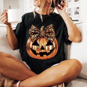 May include: Black t-shirt with a glittery orange pumpkin with a leopard print bow. The pumpkin has a jack-o'-lantern face.