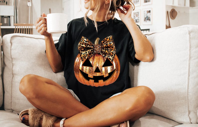 May include: Black t-shirt with a glittery orange pumpkin with a leopard print bow. The pumpkin has a jack-o'-lantern face.
