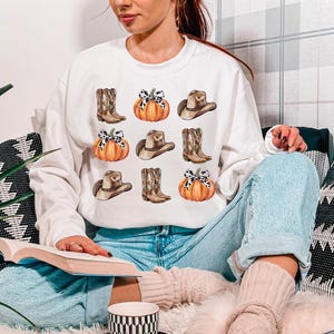 May include: White sweatshirt with a graphic print of pumpkins, cowboy hats, and boots. The graphic is in a watercolor style and features a brown, orange, and white color scheme.