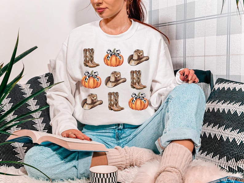 May include: White sweatshirt with a graphic print of pumpkins, cowboy hats, and boots. The graphic is in a watercolor style and features a brown, orange, and white color scheme.
