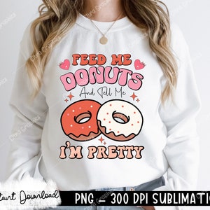 May include: White sweatshirt with a pink and red graphic design that says "Feed Me Donuts And Tell Me I'm Pretty". The graphic includes two donuts, one with pink frosting and sprinkles and the other with red frosting.