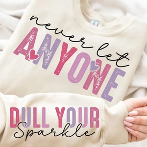 Op de afbeelding: Een crèmekleurig sweatshirt met de tekst "never let anyone dull your sparkle" in roze, paarse en zwarte letters. De tekst is verweerd en heeft een hartvormige afbeelding.