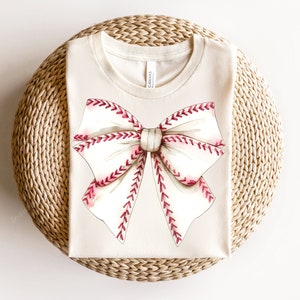 May include: A beige t-shirt with a white and red baseball-stitched bow design.