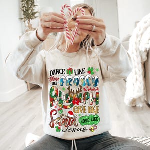 Op de afbeelding: Een wit kerst-sweatshirt met kleurrijke tekst die "Dance like Frosty, Shine like Rudolph, Give like Santa, Love like Jesus" zegt.