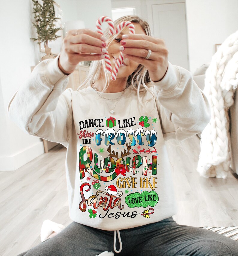 Op de afbeelding: Een wit kerst-sweatshirt met kleurrijke tekst die "Dance like Frosty, Shine like Rudolph, Give like Santa, Love like Jesus" zegt.
