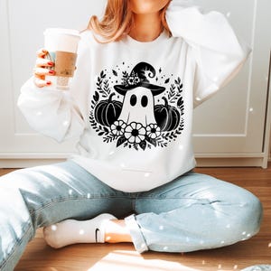 Puede incluir: Sudadera blanca con un gr&aacute;fico en blanco y negro de un fantasma con un sombrero de bruja y rodeado de calabazas y flores.