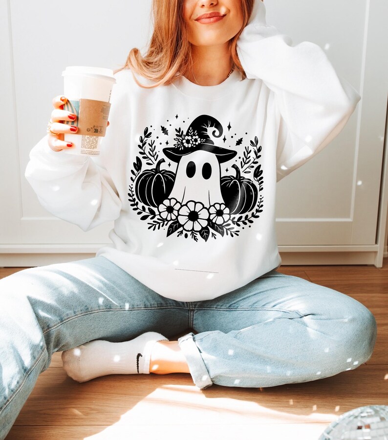 Puede incluir: Sudadera blanca con un gr&aacute;fico en blanco y negro de un fantasma con un sombrero de bruja y rodeado de calabazas y flores.