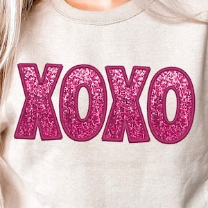 May include: A beige sweatshirt with a pink sparkly "XOXO" design.