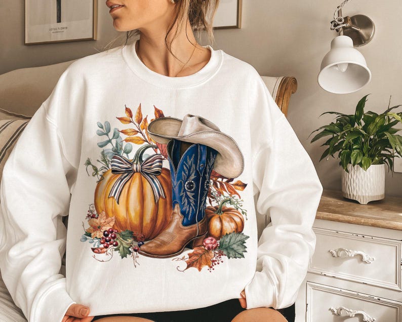 May include: White sweatshirt with a graphic print of a cowboy boot, pumpkins, and fall leaves. The boot is blue and white with a brown leather top. The pumpkins are orange and the leaves are red, orange, and yellow.