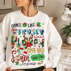 Op de afbeelding: Witte kersttrui met de tekst "Dance like Frosty, Shine like Rudolph, Give like Santa, Love like Jesus". De tekst wordt omringd door feestelijke illustraties van sneeuwvlokken, hulst, rendieren en een cadeaudoos.