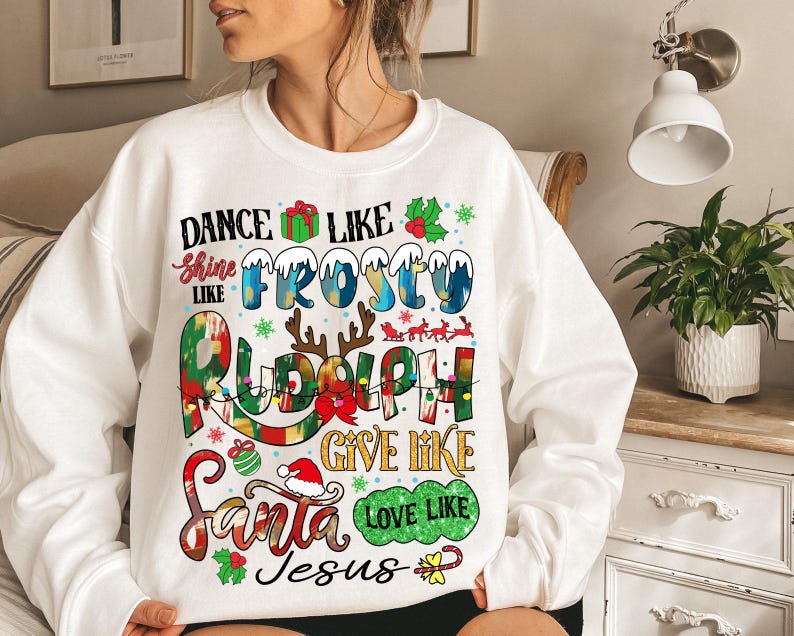 Op de afbeelding: Witte kersttrui met de tekst "Dance like Frosty, Shine like Rudolph, Give like Santa, Love like Jesus". De tekst wordt omringd door feestelijke illustraties van sneeuwvlokken, hulst, rendieren en een cadeaudoos.