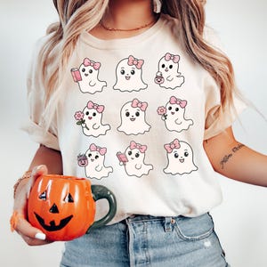 May include: A white t-shirt with a graphic of nine cute cartoon ghosts wearing pink bows. Each ghost has a different activity, such as reading a book, holding a flower, or carrying a pumpkin.