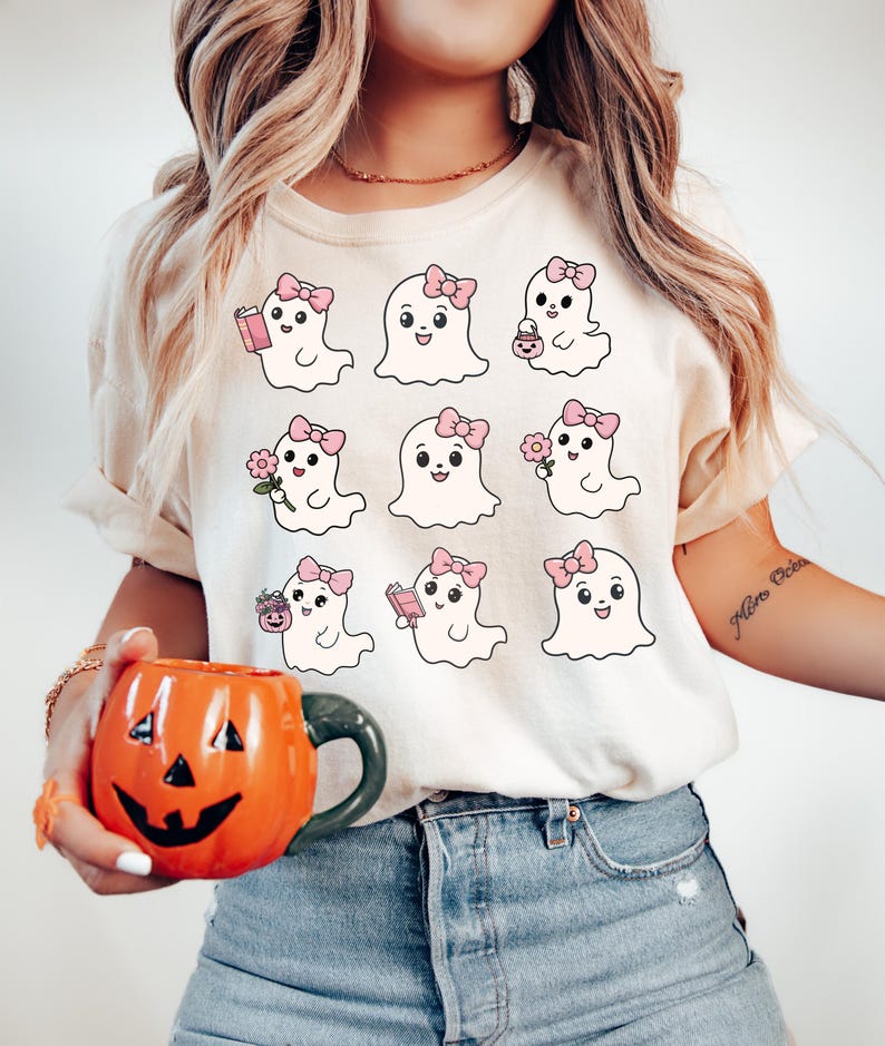 May include: A white t-shirt with a graphic of nine cute cartoon ghosts wearing pink bows. Each ghost has a different activity, such as reading a book, holding a flower, or carrying a pumpkin.