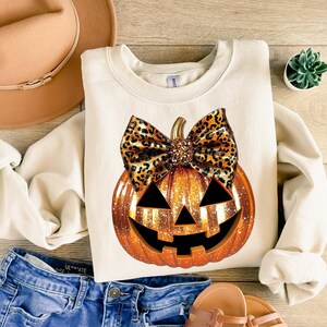 May include: A white sweatshirt with a glittery orange pumpkin with a brown and black leopard print bow. The pumpkin has a jack-o-lantern face.