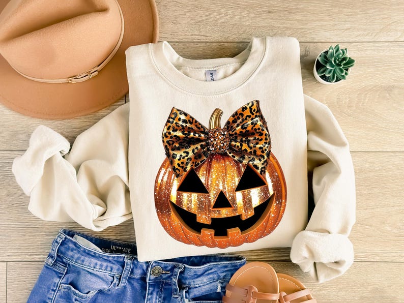 May include: A white sweatshirt with a glittery orange pumpkin with a brown and black leopard print bow. The pumpkin has a jack-o-lantern face.