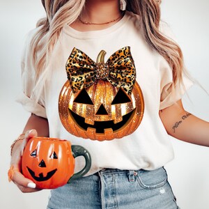 May include: A white t-shirt with a gold glitter pumpkin with a leopard print bow. The pumpkin has a jack-o-lantern face.