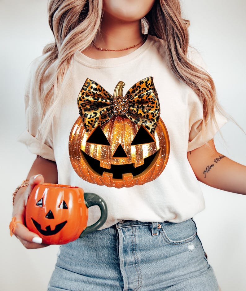 May include: A white t-shirt with a gold glitter pumpkin with a leopard print bow. The pumpkin has a jack-o-lantern face.