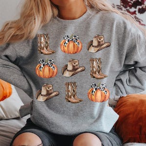May include: A gray sweatshirt with a fall-themed design featuring pumpkins, cowboy hats, and boots. The pumpkins are orange with brown stems and have a white and brown patterned bow on top. The cowboy hats are brown with a white band and the boots are brown with white accents.