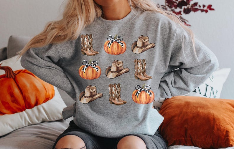 May include: A gray sweatshirt with a fall-themed design featuring pumpkins, cowboy hats, and boots. The pumpkins are orange with brown stems and have a white and brown patterned bow on top. The cowboy hats are brown with a white band and the boots are brown with white accents.