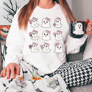 May include: White sweatshirt with a graphic of nine cute ghosts with pink bows and different accessories. The ghosts are holding a book, a flower, a cup of coffee, and a candy bag.