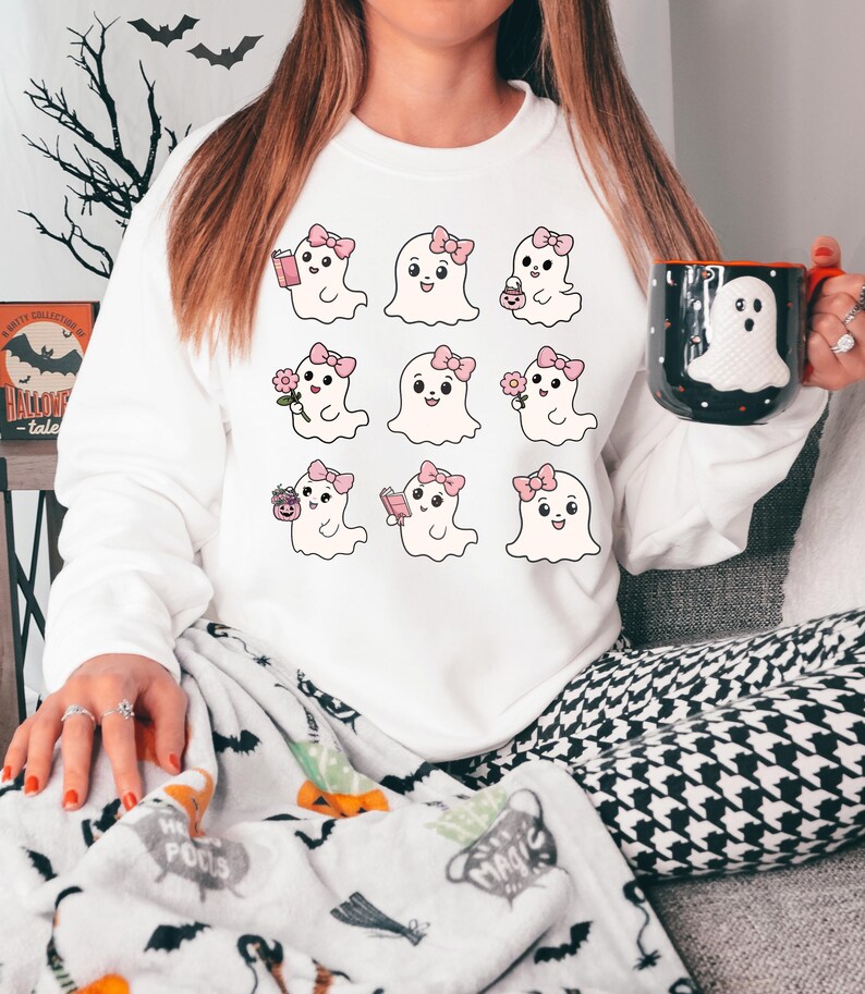 May include: White sweatshirt with a graphic of nine cute ghosts with pink bows and different accessories. The ghosts are holding a book, a flower, a cup of coffee, and a candy bag.