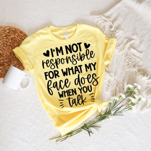 May include: A pale yellow t-shirt featuring the black text: "I'M NOT responsible FOR WHAT MY face DOES WHEN YOU talk". The shirt is folded. A white mug and decorative items are in the background.