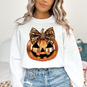 May include: White sweatshirt with a glittery orange pumpkin with a black and brown leopard print bow. The pumpkin has a jack-o-lantern face.