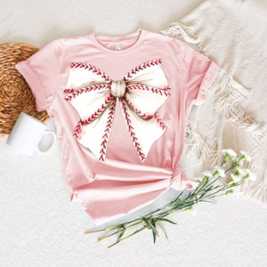 May include: A pink t-shirt with a white and red baseball-themed bow design.
