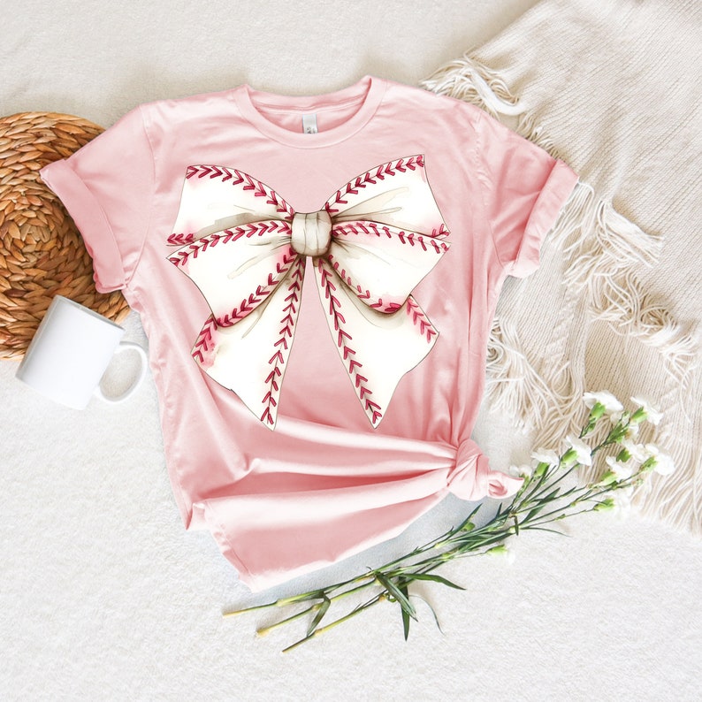 May include: A pink t-shirt with a white and red baseball-themed bow design.