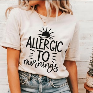 May include: A cream-coloured t-shirt with the black text "ALLERGIC TO mornings" and a sun graphic. The shirt has short sleeves and is worn with blue jeans.