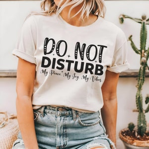 May include: A white t-shirt with the text "DO NOT DISTURB" in black lettering. The text is surrounded by small hearts and stars. Below the text is the phrase "My Peace. My Joy. My Vibes."