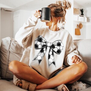 Op de afbeelding: Een cr&egrave;mekleurige sweatshirt met een groot strikdesign gemaakt van voetbalpatronen. De sweatshirt met lange mouwen wordt gedragen door iemand die op een bank zit en een zwarte mok vasthoudt.