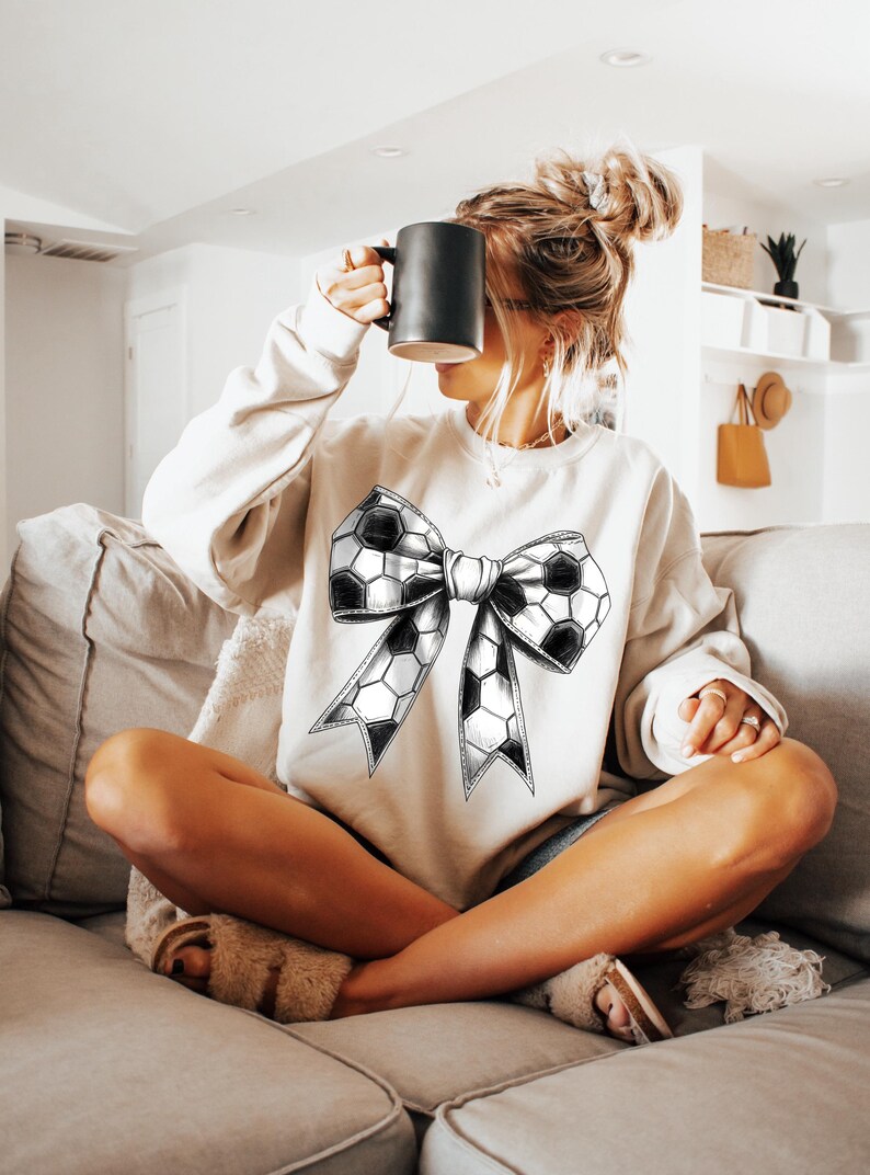 Op de afbeelding: Een cr&egrave;mekleurige sweatshirt met een groot strikdesign gemaakt van voetbalpatronen. De sweatshirt met lange mouwen wordt gedragen door iemand die op een bank zit en een zwarte mok vasthoudt.