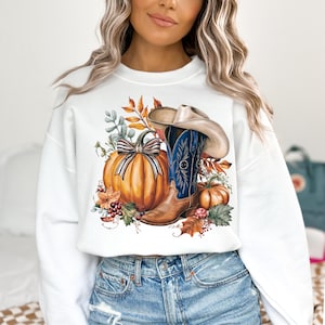 May include: White sweatshirt with a graphic print of a cowboy boot, a pumpkin, and fall leaves. The boot is blue and brown with a cowboy hat on top. The pumpkin is orange with a brown bow.