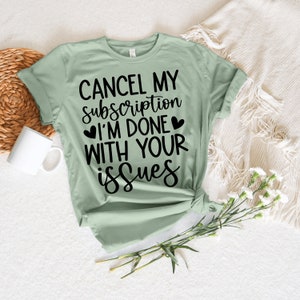 May include: A pale green t-shirt featuring the black text "CANCEL MY subscription I'M DONE WITH YOUR issues". The shirt is knotted at the hem. A white mug and flowers are in the background.