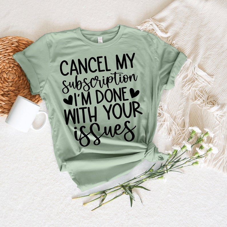 May include: A pale green t-shirt featuring the black text "CANCEL MY subscription I'M DONE WITH YOUR issues". The shirt is knotted at the hem. A white mug and flowers are in the background.