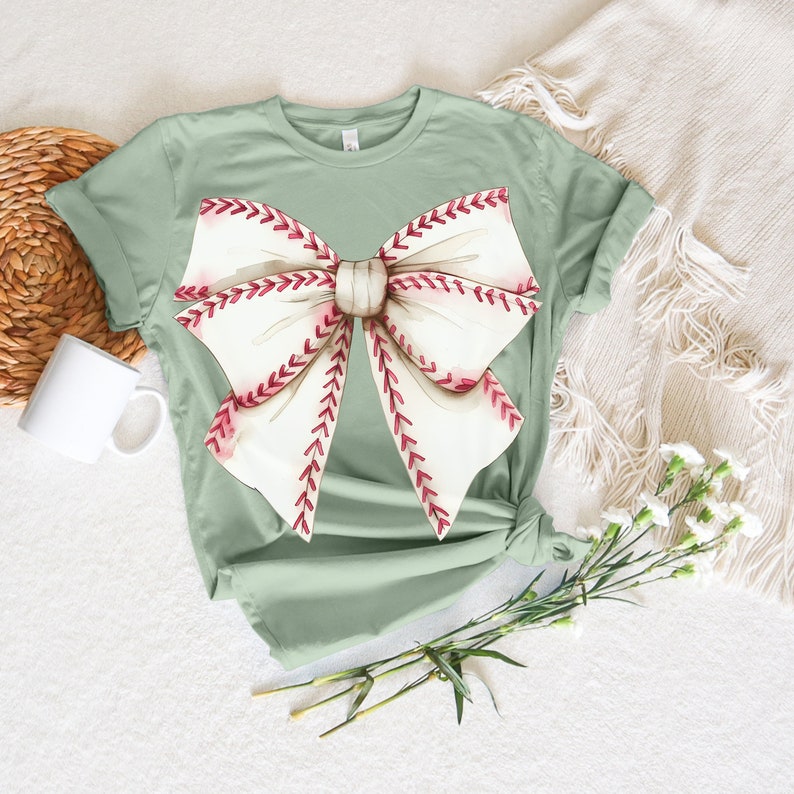 May include: A sage green t-shirt with a white bow design. The bow is made of baseball stitching with red stitching.