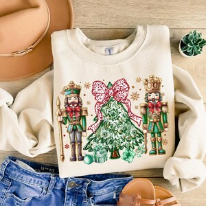 May include: A cream-colored sweatshirt with a Christmas tree design featuring two nutcracker figures, a red bow, and gold accents.
