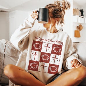 Puede incluir: Una sudadera blanca con un diseño rojo y blanco. El diseño presenta una corona de espinas, una cruz y el texto "the original love letters".