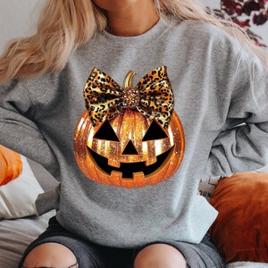 May include: A gray sweatshirt with a glittery orange pumpkin with a leopard print bow. The pumpkin has a jack-o-lantern face.