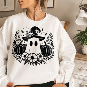 Puede incluir: Sudadera blanca con un gr&aacute;fico en blanco y negro de un fantasma con un sombrero de bruja y sosteniendo calabazas. El gr&aacute;fico est&aacute; rodeado de hojas y flores negras.