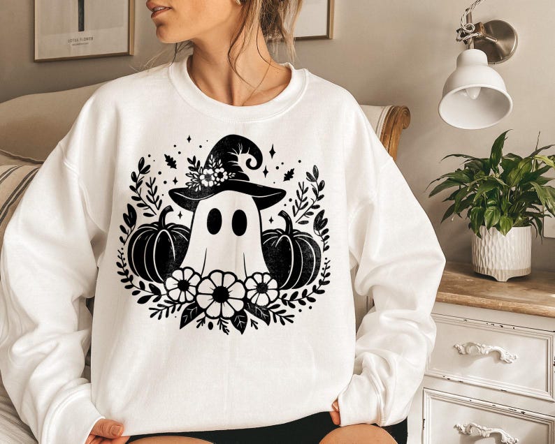 Puede incluir: Sudadera blanca con un gr&aacute;fico en blanco y negro de un fantasma con un sombrero de bruja y sosteniendo calabazas. El gr&aacute;fico est&aacute; rodeado de hojas y flores negras.