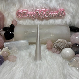 May include: A white cone-shaped crystal point on a white fluffy surface surrounded by various crystals and stones. The crystals are pink, white, and black. The text "@EveryMySparkle" is visible in the background.