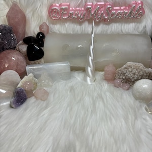 May include: A collection of pink, white, and purple crystals and stones, including a unicorn horn, on a white fluffy surface. The text "@EnvyMySparkle" is displayed on a pink sign.