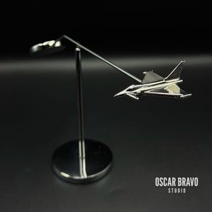 Può includere: Un modello di aereo in metallo argentato lucido montato su un supporto. Il modello ha ali dettagliate e un design elegante. Il supporto ha una base circolare. Il testo "OSCAR BRAVO STUDIO" è visibile.