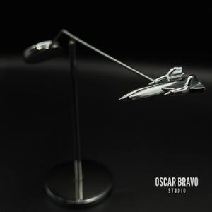 Può includere: Una scultura in metallo argentato lucido di un aereo a reazione, montata su un supporto sottile con una base circolare. La scultura è angolata, dando l'impressione di volo. Lo sfondo è nero pieno, con il testo "OSCAR BRAVO STUDIO" in bianco.