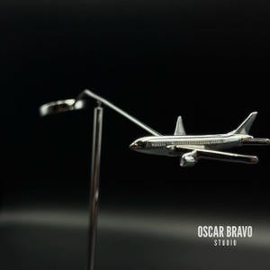 May include: A silver-colored airplane model with detailed wings and engines is suspended in mid-air by a thin metal rod. The model is set against a dark background. The text "OSCAR BRAVO STUDIO" is visible.