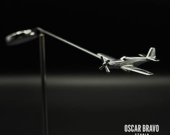 Könnte beinhalten: Ein silbernes Flugzeugmodell mit Propeller, montiert auf einer dünnen Metallstange. Das Modell hat Flügel, ein Heck und ein Cockpit. Der Sockel ist ein kleiner, runder, schwarzer Ständer. Der Text "OSCAR BRAVO STUDIO" steht unten.