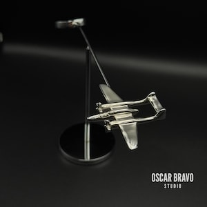May include: A silver airplane sculpture, intricately designed and mounted on a slender metal stand with a circular base. The sculpture is set against a dark backdrop, with the text "OSCAR BRAVO STUDIO" visible.