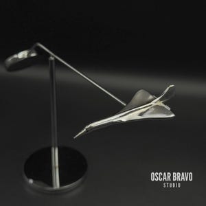 Könnte beinhalten: Eine silberfarbene Concorde-Modellflugzeugskulptur auf einem Ständer. Die Skulptur ist sehr detailliert und reflektiert das Licht, mit einem schlanken, aerodynamischen Design. Der Ständer ist aus Metall und hat einen kreisförmigen Sockel. Der Text "OSCAR BRAVO STUDIO" ist sichtbar.